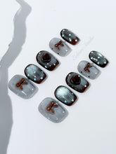 Short coffin press-on nails in mint and chocolate brown gradient with 3D heart and bow accents, displayed in a clear Éclaire Studio case under soft lighting.