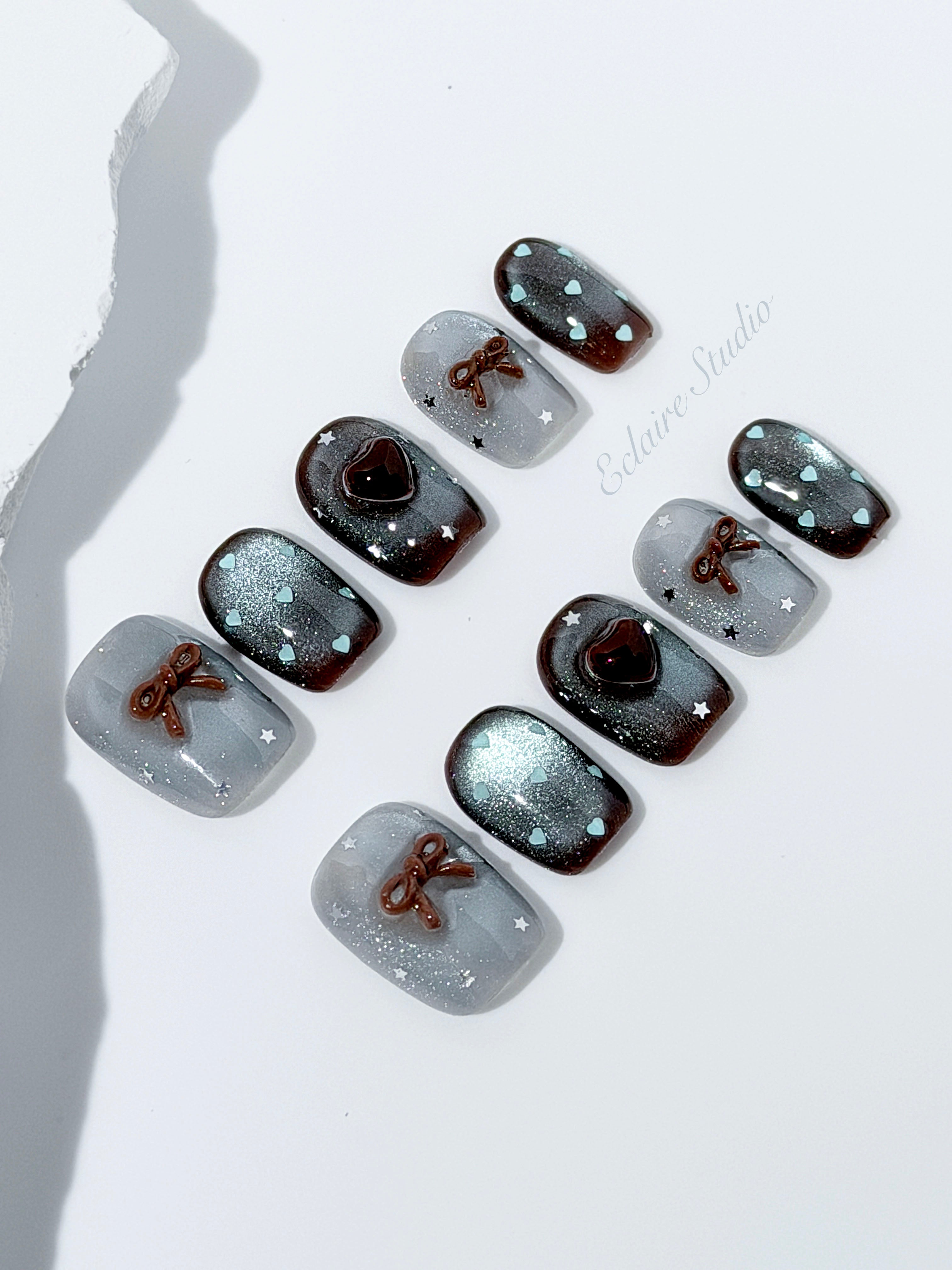 Short coffin press-on nails in mint and chocolate brown gradient with 3D heart and bow accents, displayed in a clear Éclaire Studio case under soft lighting.