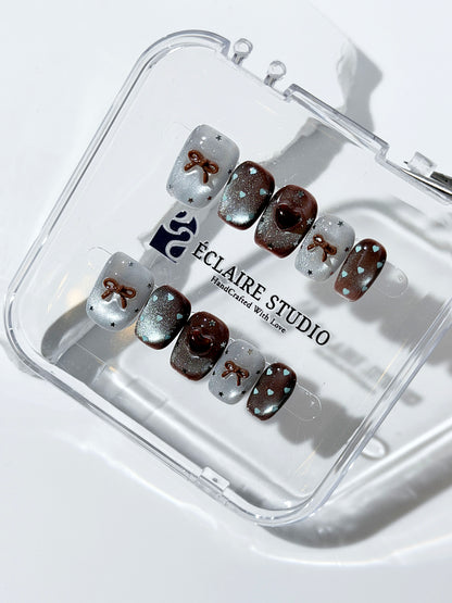 Short coffin press-on nails in mint and chocolate brown gradient with 3D heart and bow accents, displayed in a clear Éclaire Studio case under soft lighting.