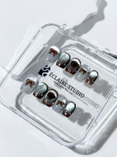 Short coffin press-on nails in mint and chocolate brown gradient with 3D heart and bow accents, displayed in a clear Éclaire Studio case under soft lighting.