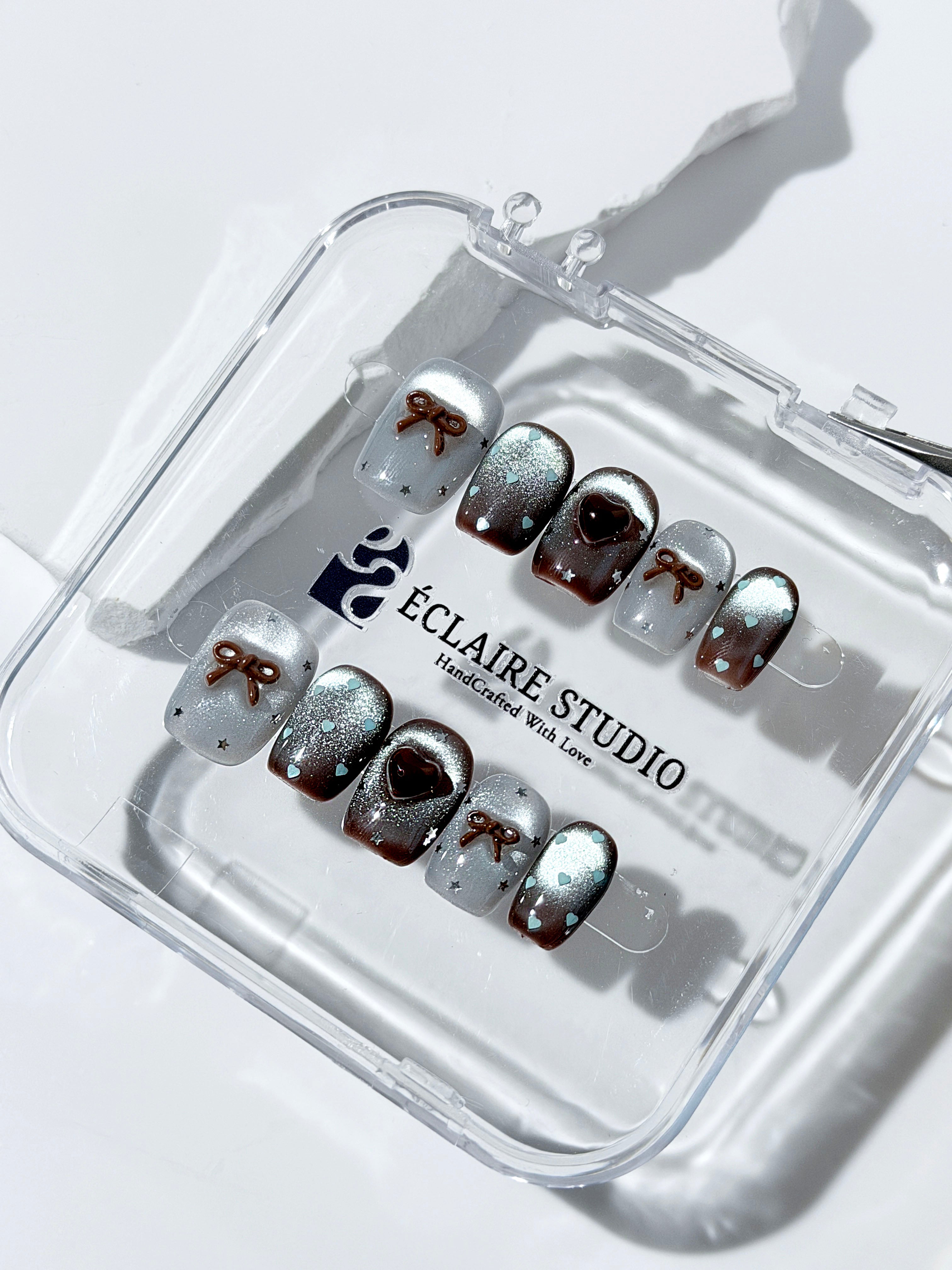 Short coffin press-on nails in mint and chocolate brown gradient with 3D heart and bow accents, displayed in a clear Éclaire Studio case under soft lighting.