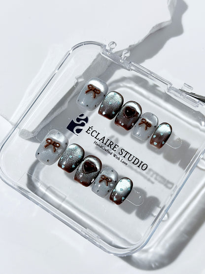 Short coffin press-on nails in mint and chocolate brown gradient with 3D heart and bow accents, displayed in a clear Éclaire Studio case under soft lighting.