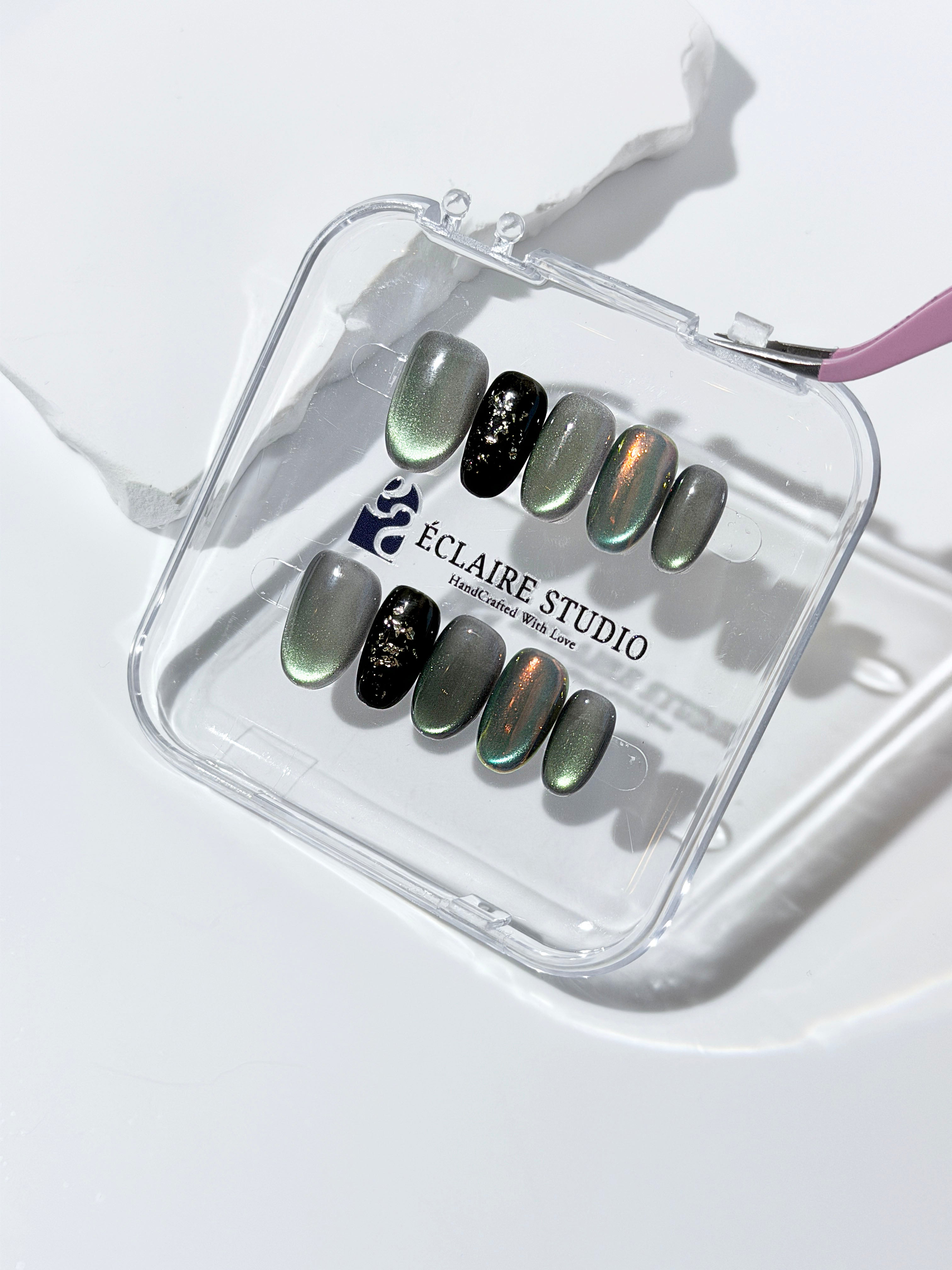 Short round press-on nails in shifting evergreen cat-eye shimmer with black and gold foil accent, displayed in a clear Éclaire Studio case.