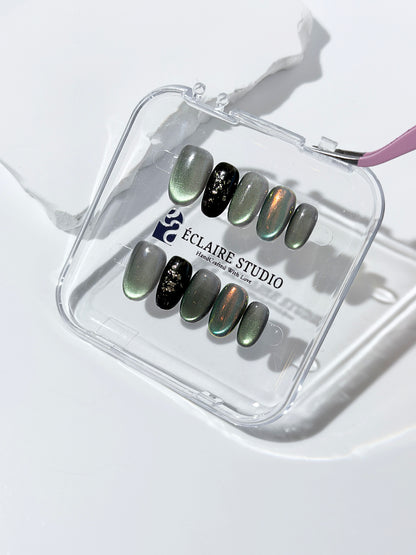 Short round press-on nails in shifting evergreen cat-eye shimmer with black and gold foil accent, displayed in a clear Éclaire Studio case.