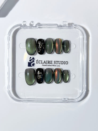 Short round press-on nails in shifting evergreen cat-eye shimmer with black and gold foil accent, displayed in a clear Éclaire Studio case.