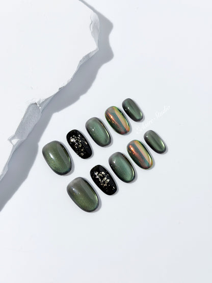 Short round press-on nails in shifting evergreen cat-eye shimmer with black and gold foil accent, displayed in a clear Éclaire Studio case.