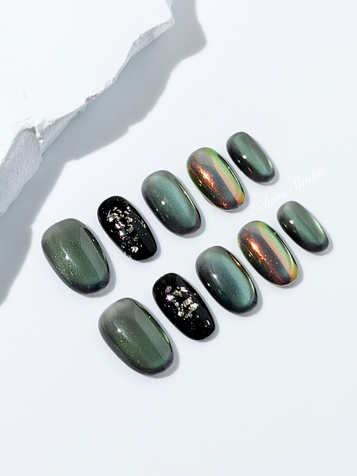 Short round press-on nails in shifting evergreen cat-eye shimmer with black and gold foil accent, displayed in a clear Éclaire Studio case.
