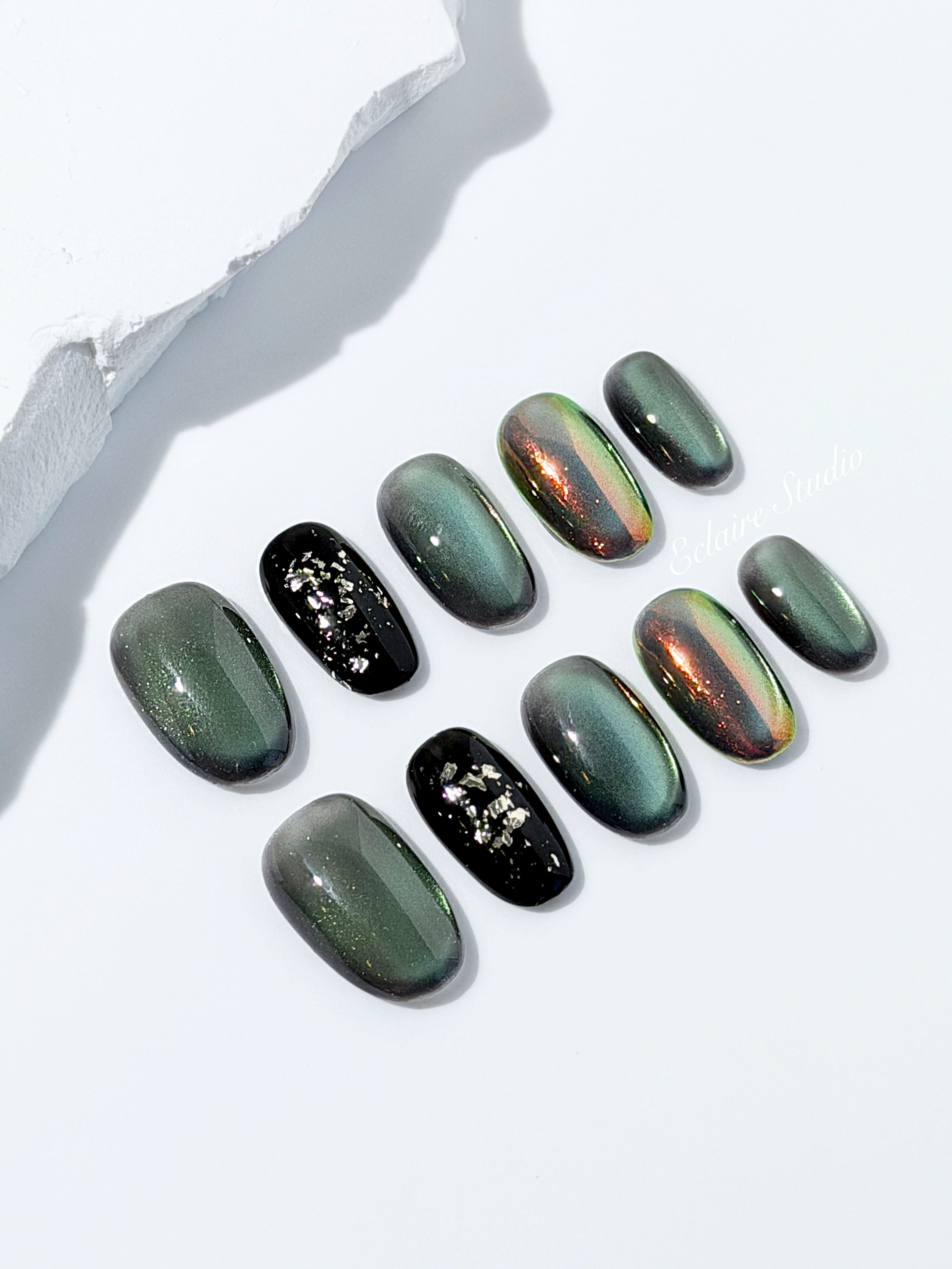 Short round press-on nails in shifting evergreen cat-eye shimmer with black and gold foil accent, displayed in a clear Éclaire Studio case.