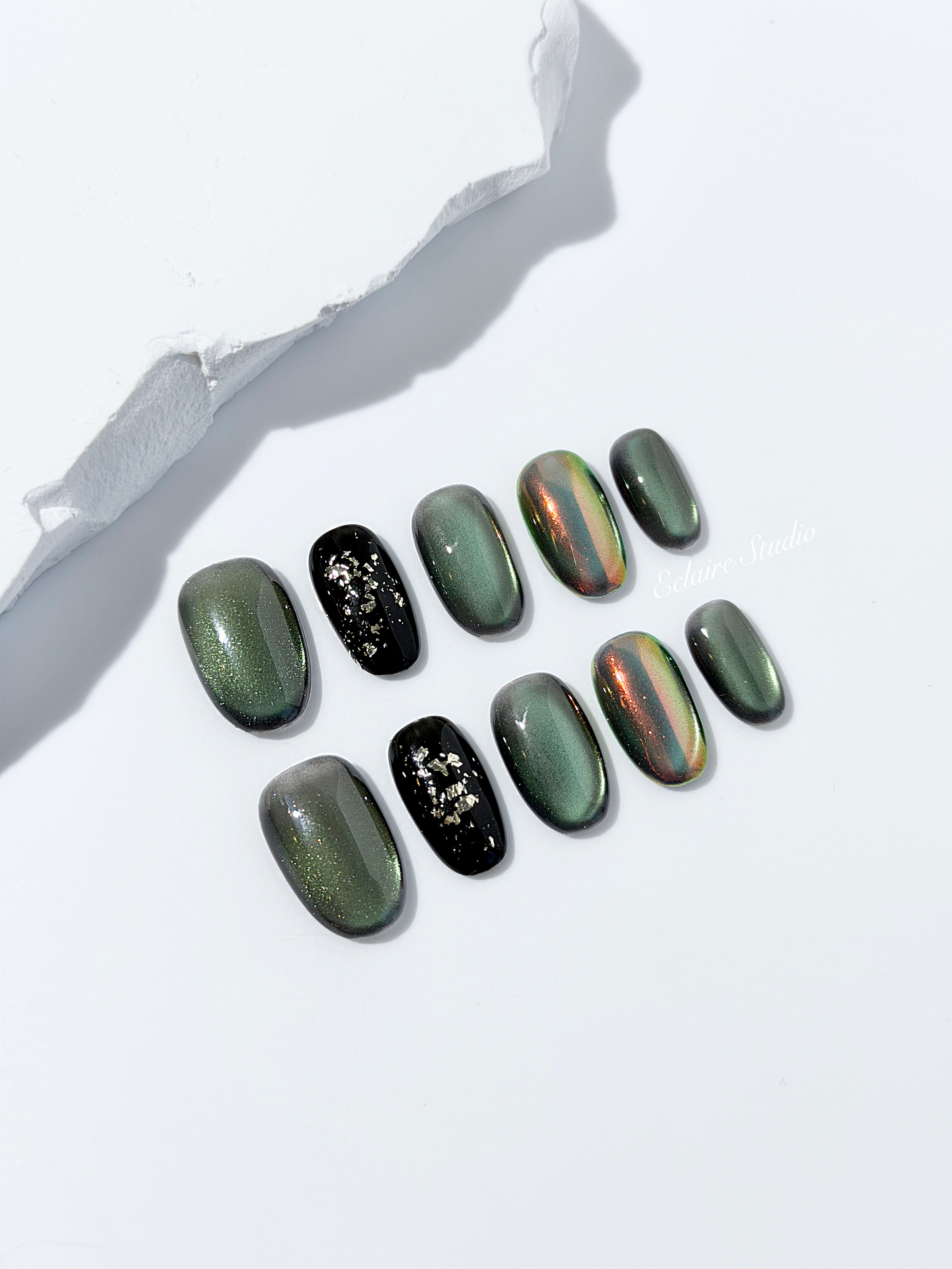 Short round press-on nails in shifting evergreen cat-eye shimmer with black and gold foil accent, displayed in a clear Éclaire Studio case.