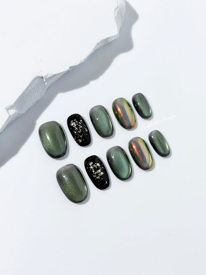 Short round press-on nails in shifting evergreen cat-eye shimmer with black and gold foil accent, displayed in a clear Éclaire Studio case.
