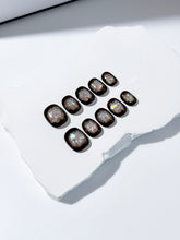 Short squoval press-on nails with dark brown base and iridescent frosted shimmer center, styled on white stone background.