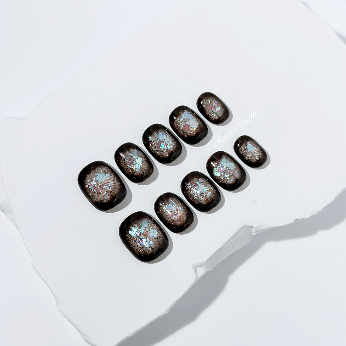 Short squoval press-on nails with dark brown base and iridescent frosted shimmer center, styled on white stone background.