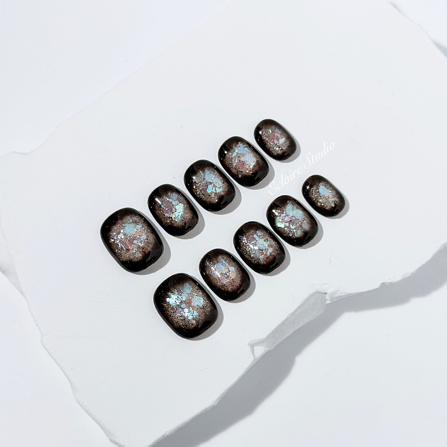 Short squoval press-on nails with dark brown base and iridescent frosted shimmer center, styled on white stone background.
