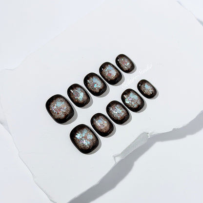 Short squoval press-on nails with dark brown base and iridescent frosted shimmer center, styled on white stone background.