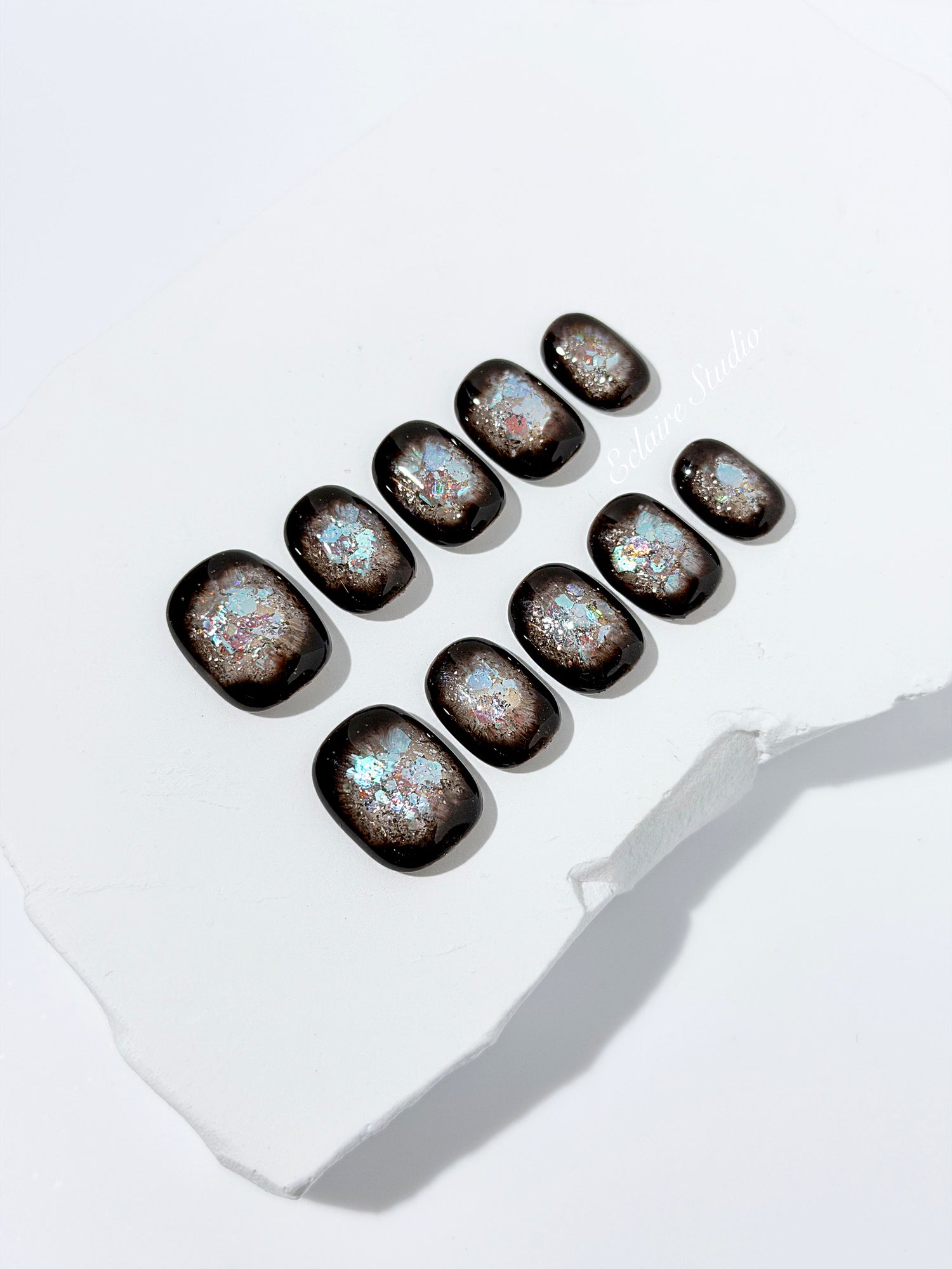Short squoval press-on nails with dark brown base and iridescent frosted shimmer center, styled on white stone background.