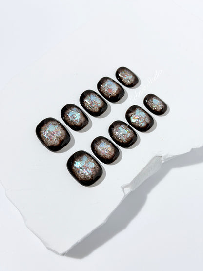 Short squoval press-on nails with dark brown base and iridescent frosted shimmer center, styled on white stone background.