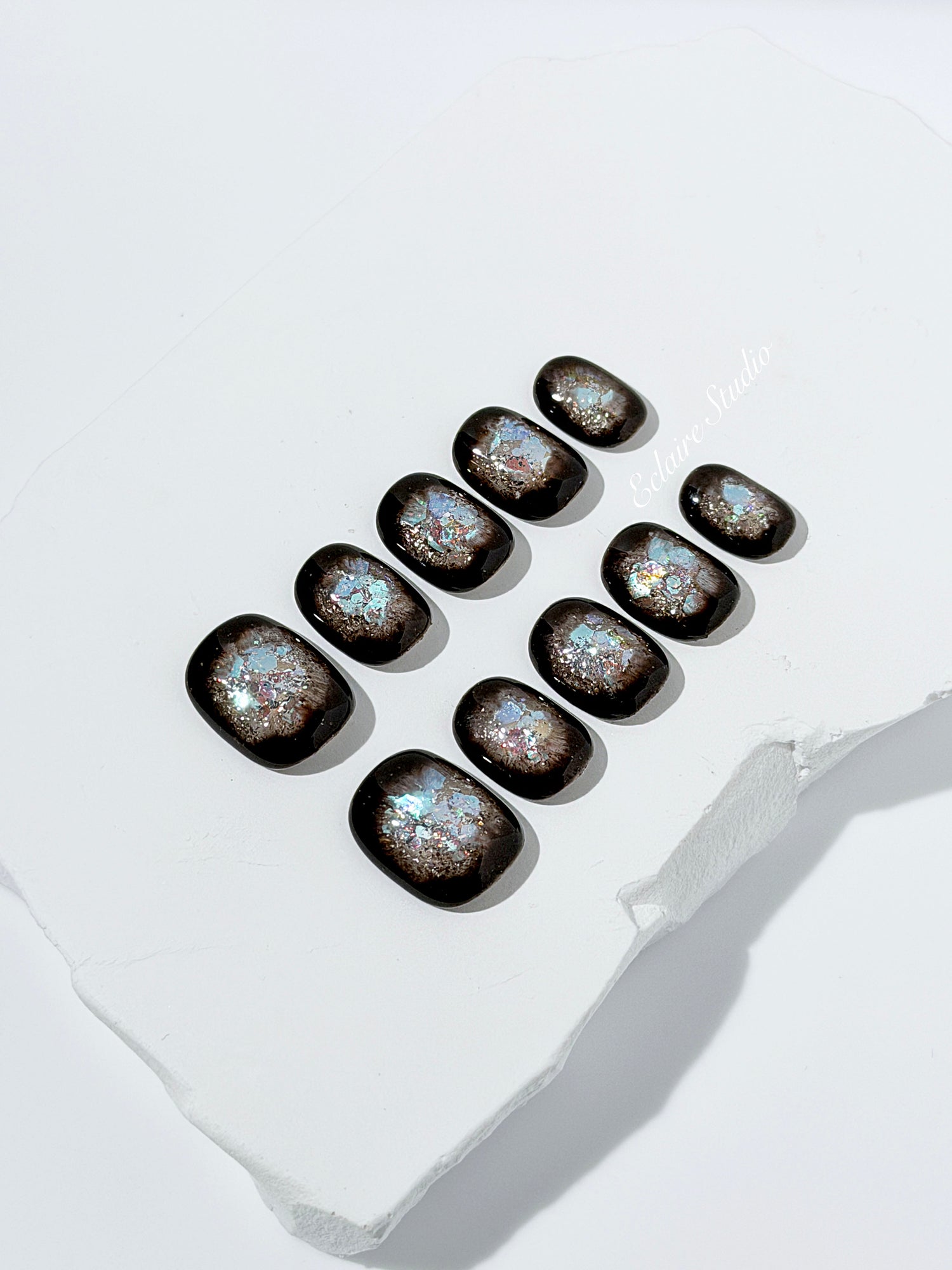 Short squoval press-on nails with dark brown base and iridescent frosted shimmer center, styled on white stone background.