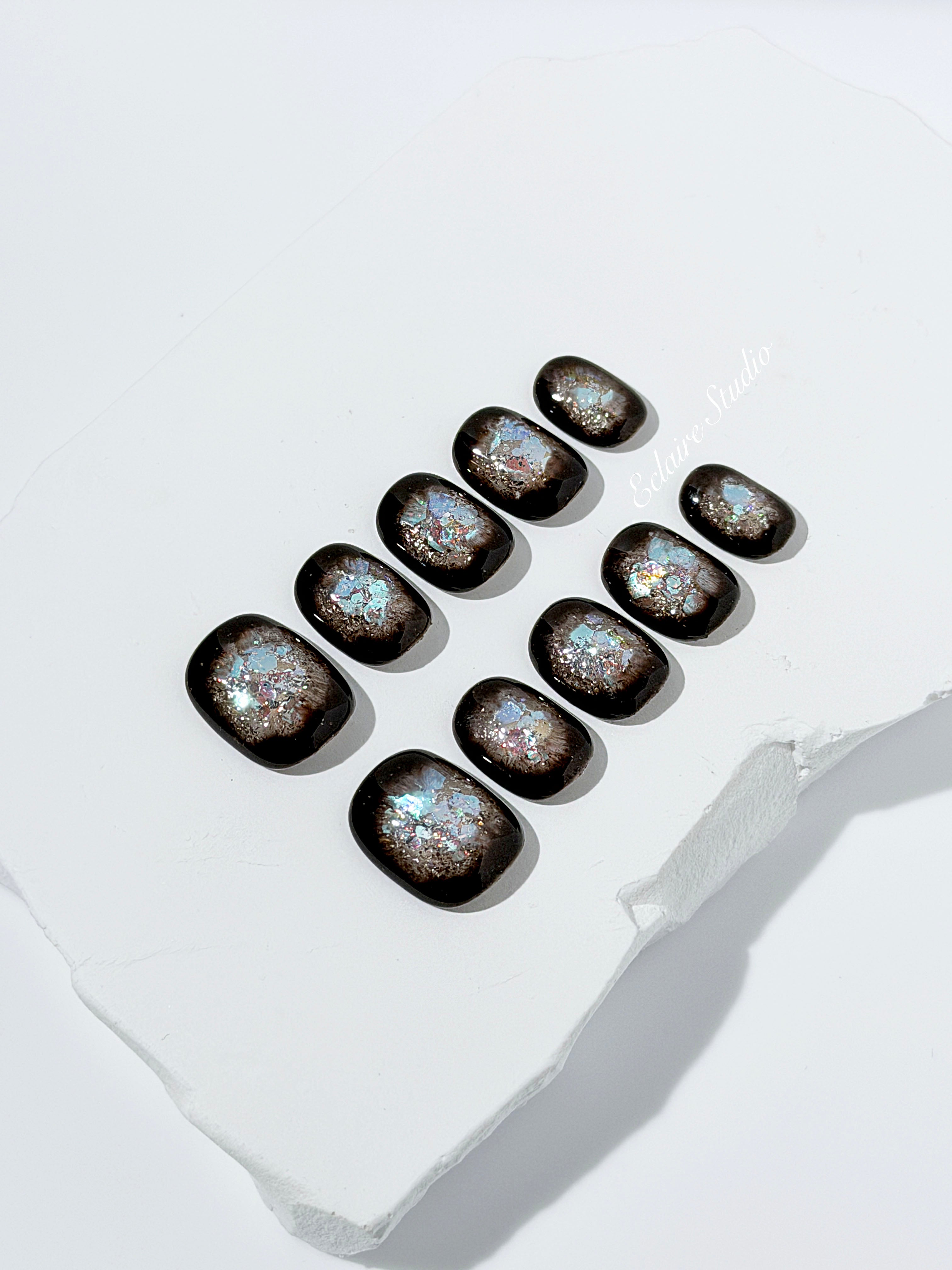 Short squoval press-on nails with dark brown base and iridescent frosted shimmer center, styled on white stone background.