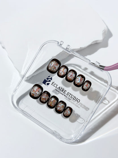 Short squoval press-on nails with dark brown base and iridescent frosted shimmer center, styled on white stone background.