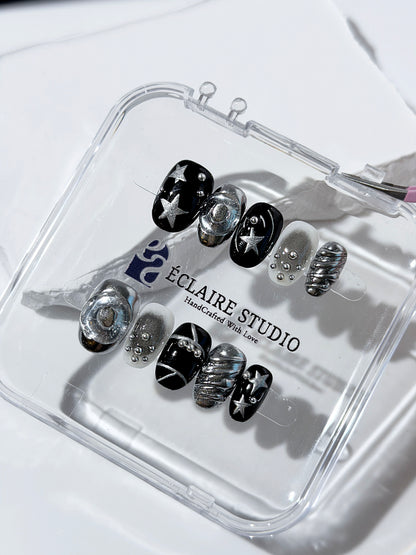 Black and silver press-on nails with 3D chrome swirl accents, star details, and shimmer gradient in a bold winter aesthetic.
