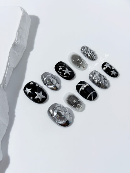 Black and silver press-on nails with 3D chrome swirl accents, star details, and shimmer gradient in a bold winter aesthetic.
