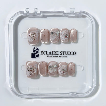 Short coffin pink nude cat-eye press-on nails with lace texture and pearl bow accents in a soft romantic winter style.
