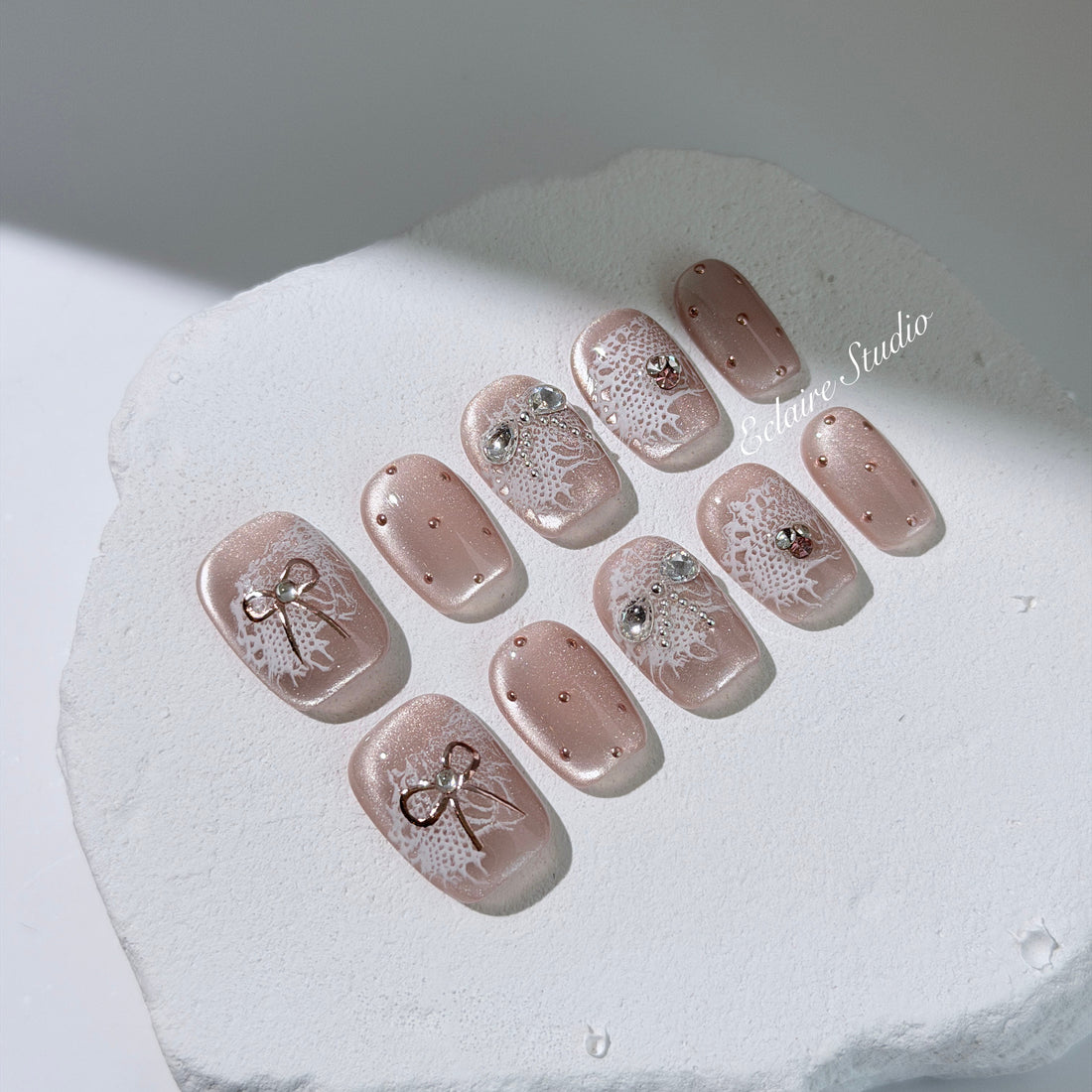 Short coffin pink nude cat-eye press-on nails with lace texture and pearl bow accents in a soft romantic winter style.
