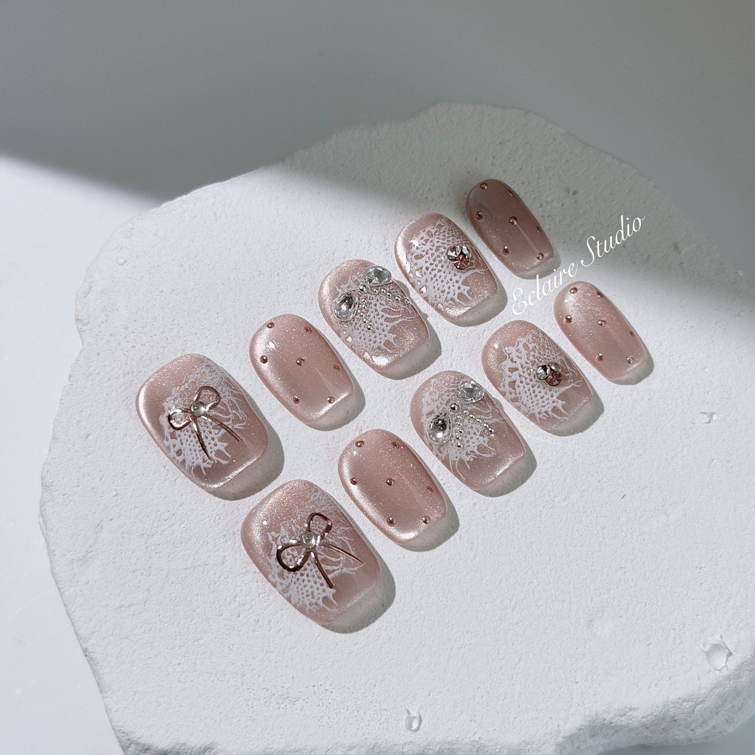 Short coffin pink nude cat-eye press-on nails with lace texture and pearl bow accents in a soft romantic winter style.
