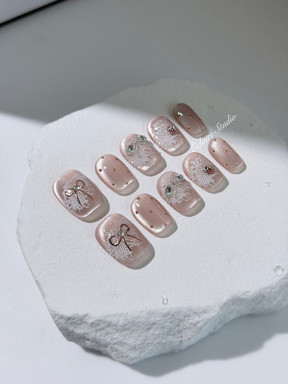 Short coffin pink nude cat-eye press-on nails with lace texture and pearl bow accents in a soft romantic winter style.
