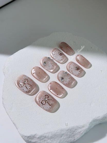 Short coffin pink nude cat-eye press-on nails with lace texture and pearl bow accents in a soft romantic winter style.

