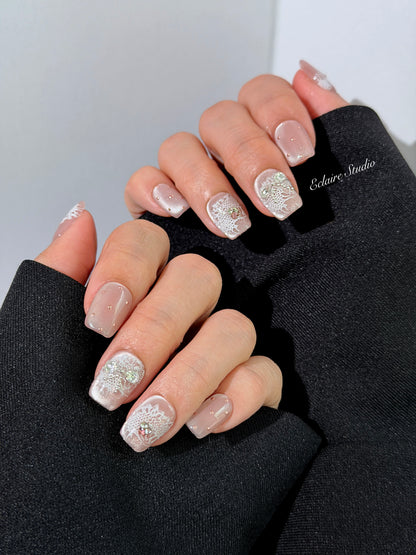 Short coffin pink nude cat-eye press-on nails with lace texture and pearl bow accents in a soft romantic winter style.
