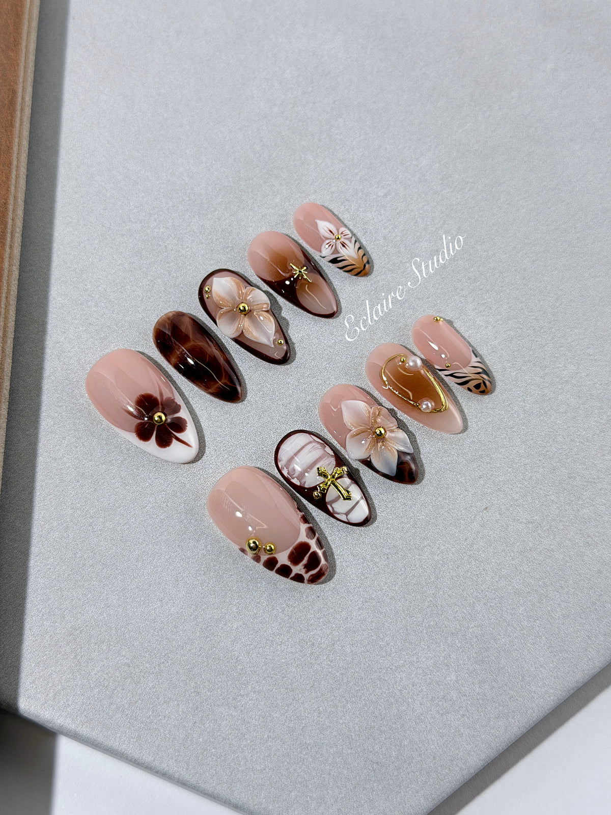 Petal Locket almond press-on nails with nude bases, ivory 3D flowers, brown florals, tortoiseshell, animal-print French tips, pearls, and gold accents.
