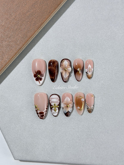 Petal Locket almond press-on nails with nude bases, ivory 3D flowers, brown florals, tortoiseshell, animal-print French tips, pearls, and gold accents.
