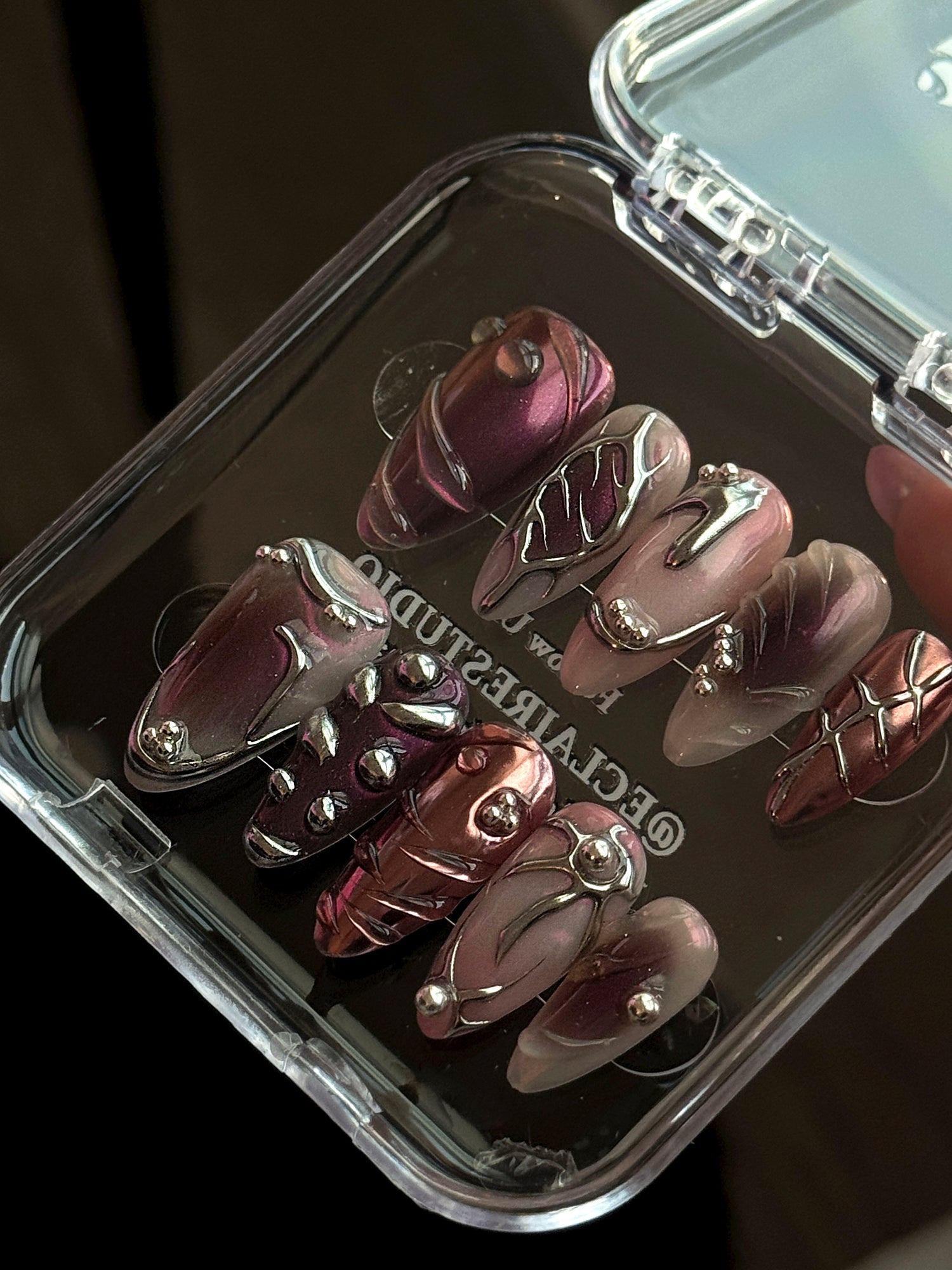 Set of long, almond-shaped press-on nails with a pink-to-purple multichrome base, accented by sculpted chrome designs, metallic textures, and small silver studs, displayed on a ribbed reflective surface under natural light.
