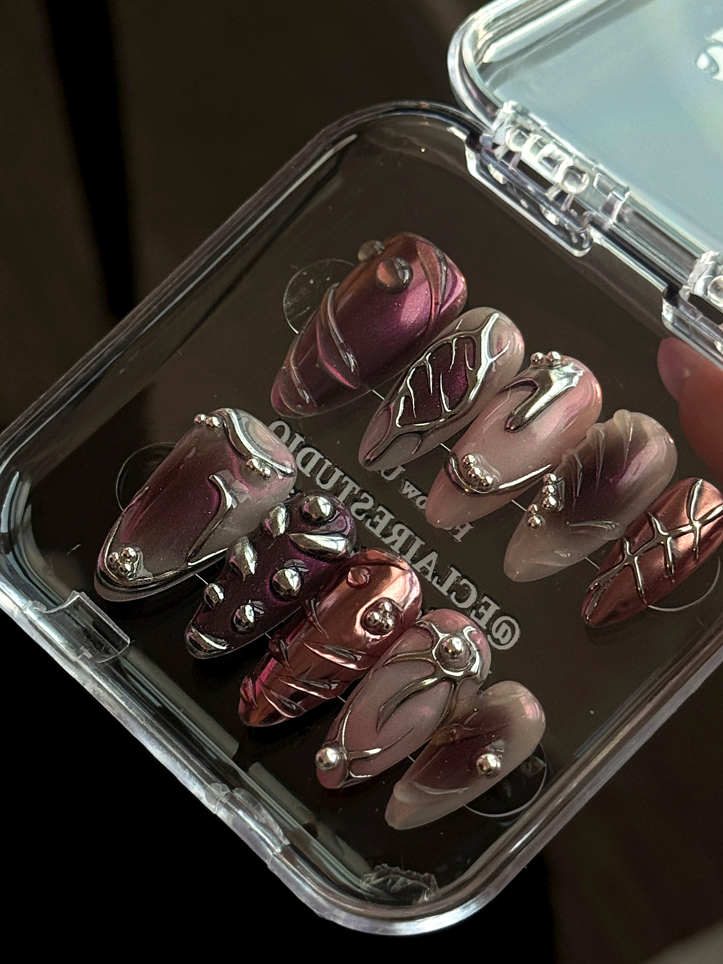 Set of long, almond-shaped press-on nails with a pink-to-purple multichrome base, accented by sculpted chrome designs, metallic textures, and small silver studs, displayed on a ribbed reflective surface under natural light.