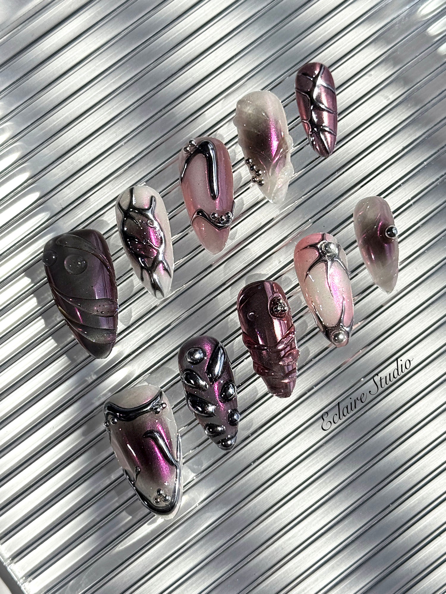 Set of long, almond-shaped press-on nails with a pink-to-purple multichrome base, accented by sculpted chrome designs, metallic textures, and small silver studs, displayed on a ribbed reflective surface under natural light.