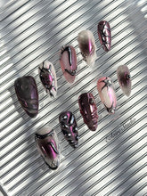 Set of long, almond-shaped press-on nails with a pink-to-purple multichrome base, accented by sculpted chrome designs, metallic textures, and small silver studs, displayed on a ribbed reflective surface under natural light.