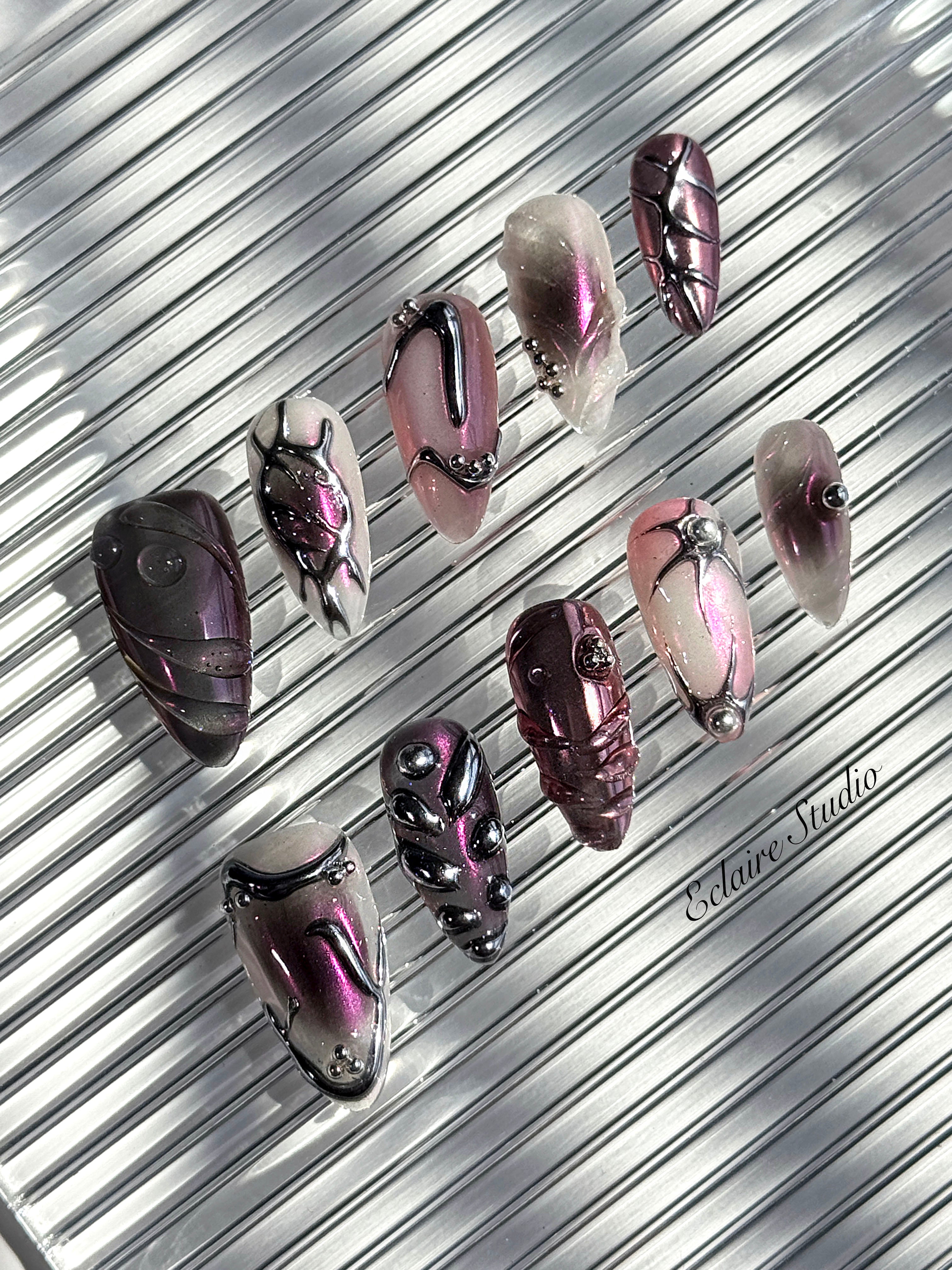 Set of long, almond-shaped press-on nails with a pink-to-purple multichrome base, accented by sculpted chrome designs, metallic textures, and small silver studs, displayed on a ribbed reflective surface under natural light.