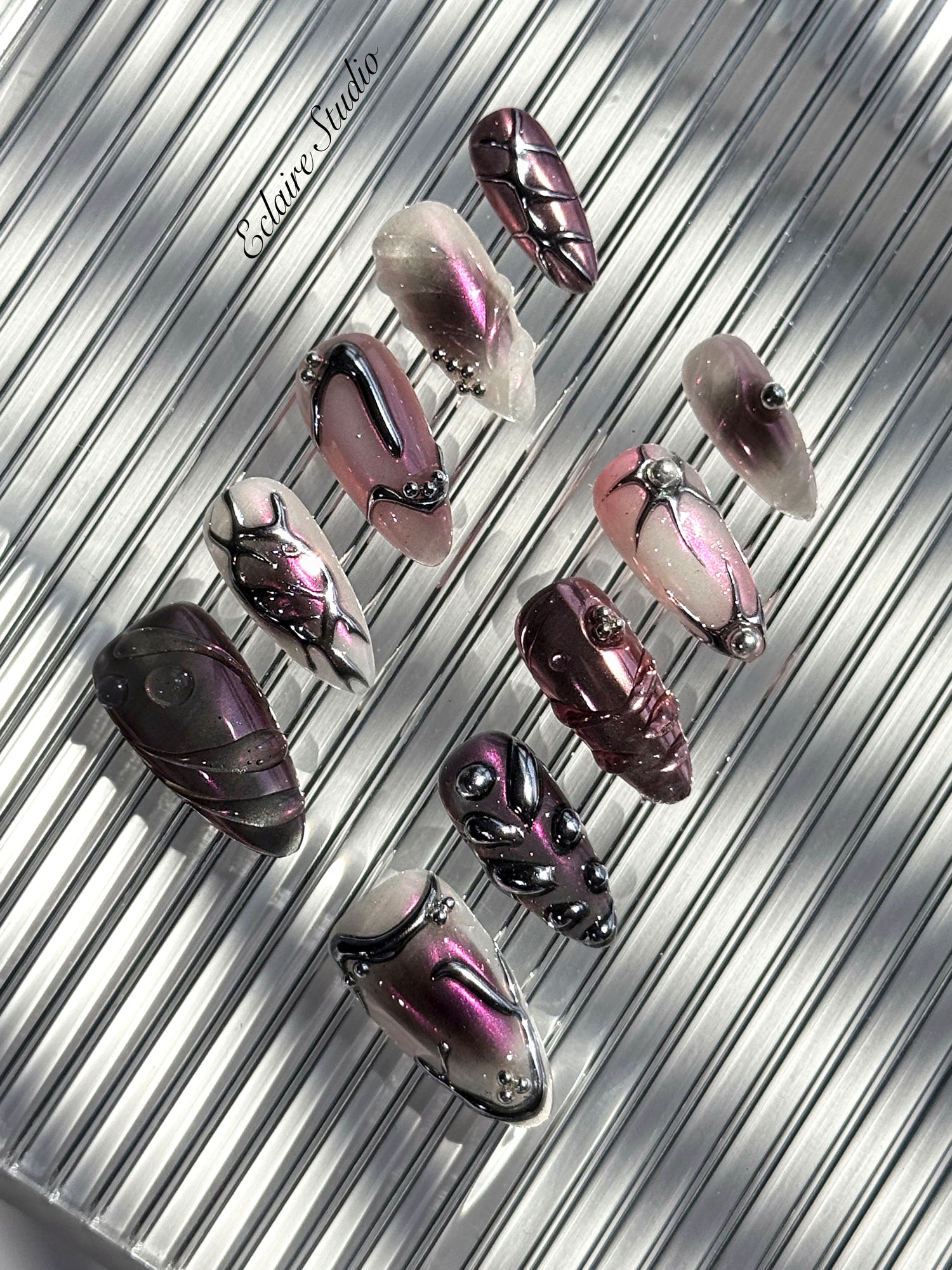 Set of long, almond-shaped press-on nails with a pink-to-purple multichrome base, accented by sculpted chrome designs, metallic textures, and small silver studs, displayed on a ribbed reflective surface under natural light.