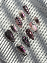 Set of long, almond-shaped press-on nails with a pink-to-purple multichrome base, accented by sculpted chrome designs, metallic textures, and small silver studs, displayed on a ribbed reflective surface under natural light.