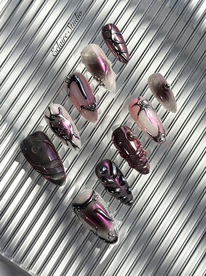 Set of long, almond-shaped press-on nails with a pink-to-purple multichrome base, accented by sculpted chrome designs, metallic textures, and small silver studs, displayed on a ribbed reflective surface under natural light.