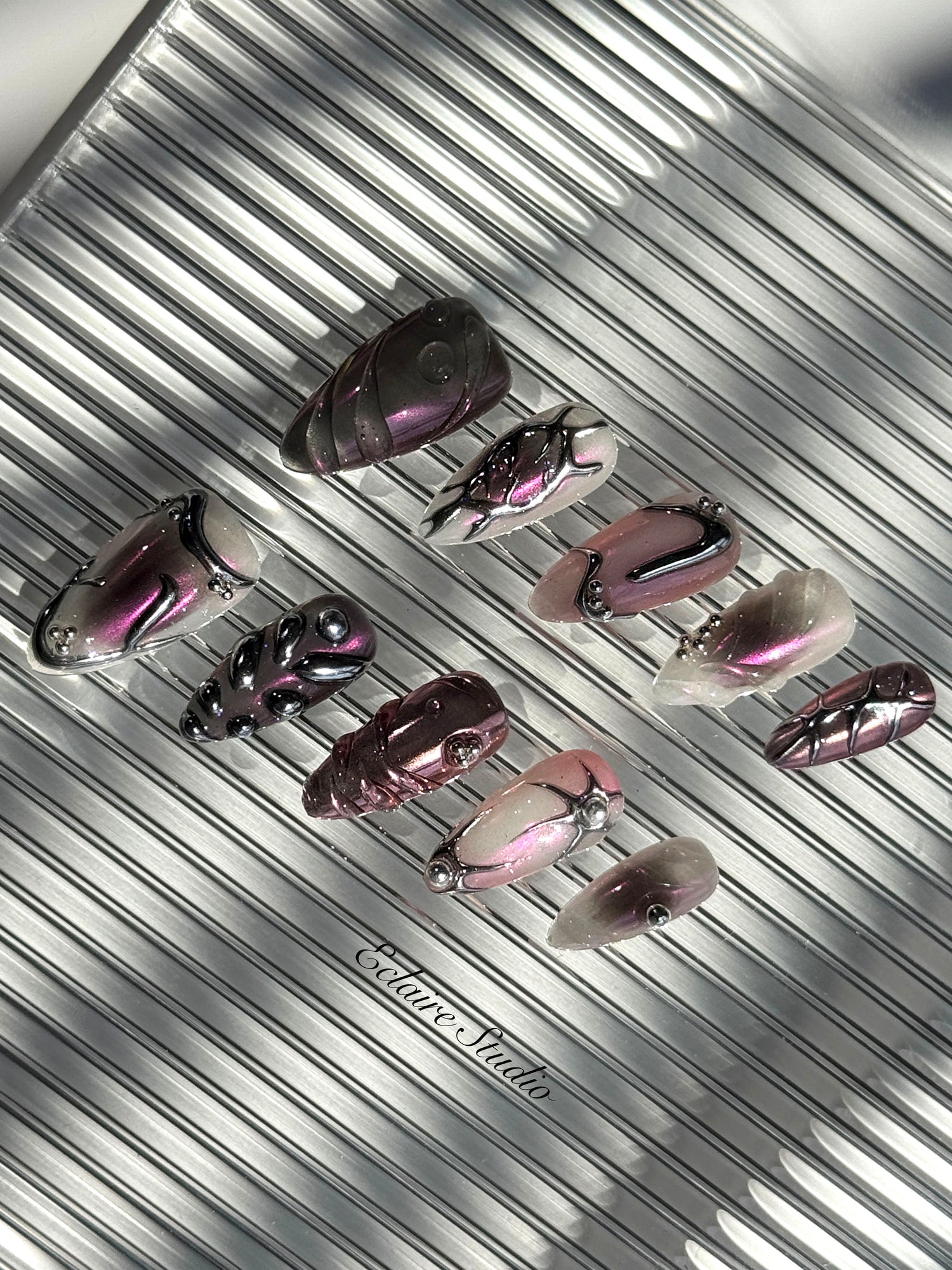 Set of long, almond-shaped press-on nails with a pink-to-purple multichrome base, accented by sculpted chrome designs, metallic textures, and small silver studs, displayed on a ribbed reflective surface under natural light.