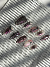 Set of long, almond-shaped press-on nails with a pink-to-purple multichrome base, accented by sculpted chrome designs, metallic textures, and small silver studs, displayed on a ribbed reflective surface under natural light.