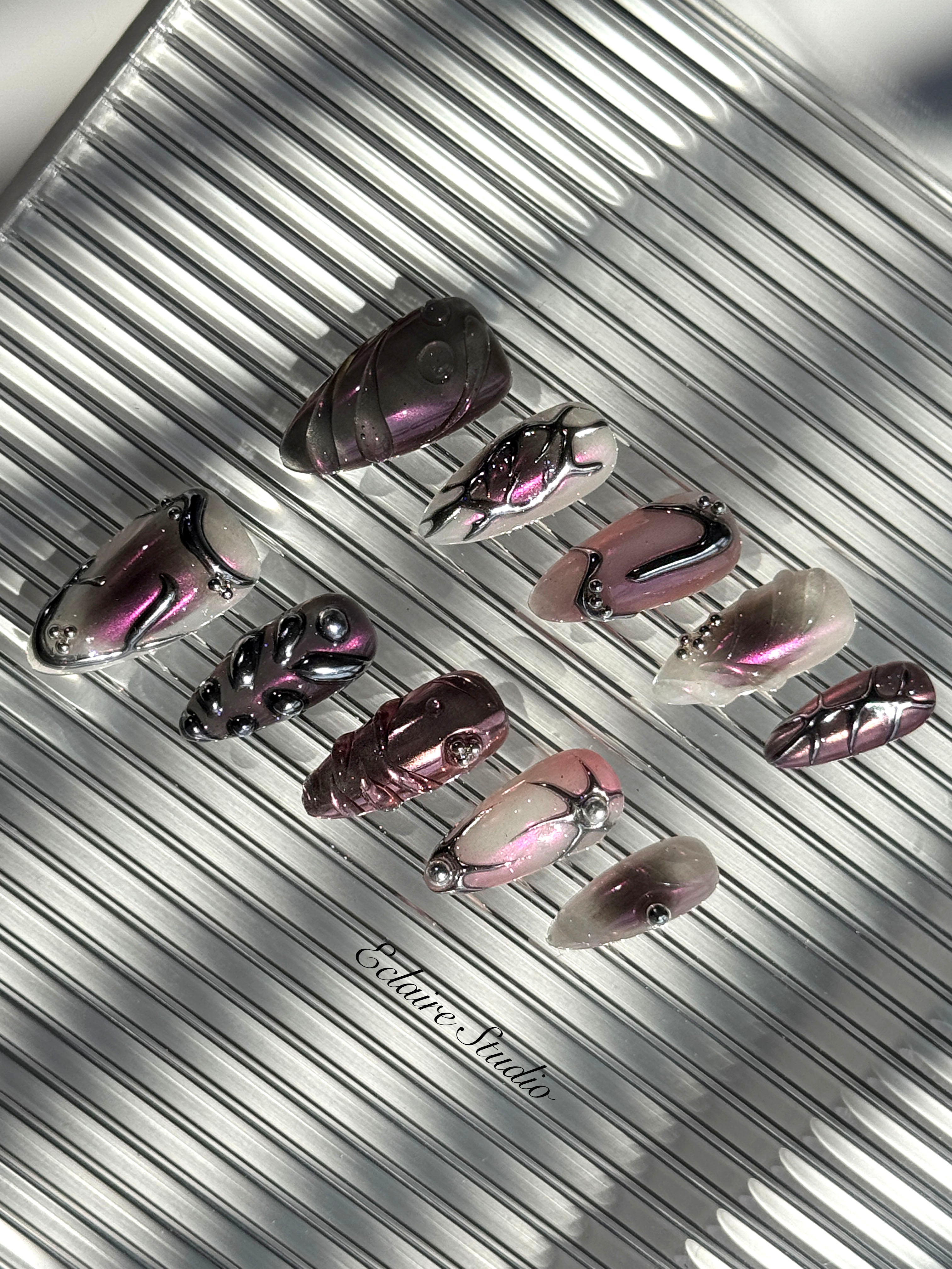 Set of long, almond-shaped press-on nails with a pink-to-purple multichrome base, accented by sculpted chrome designs, metallic textures, and small silver studs, displayed on a ribbed reflective surface under natural light.