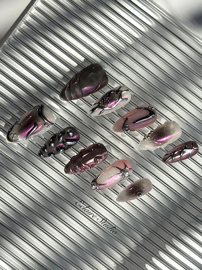 Set of long, almond-shaped press-on nails with a pink-to-purple multichrome base, accented by sculpted chrome designs, metallic textures, and small silver studs, displayed on a ribbed reflective surface under natural light.