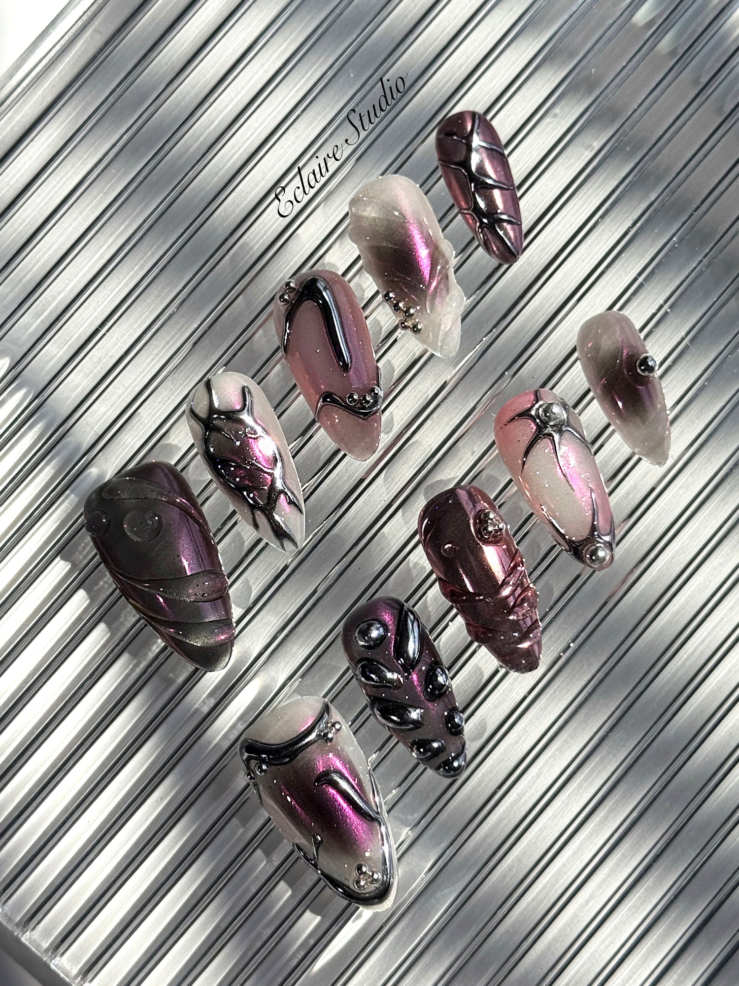 Set of long, almond-shaped press-on nails with a pink-to-purple multichrome base, accented by sculpted chrome designs, metallic textures, and small silver studs, displayed on a ribbed reflective surface under natural light.