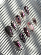 Set of long, almond-shaped press-on nails with a pink-to-purple multichrome base, accented by sculpted chrome designs, metallic textures, and small silver studs, displayed on a ribbed reflective surface under natural light.