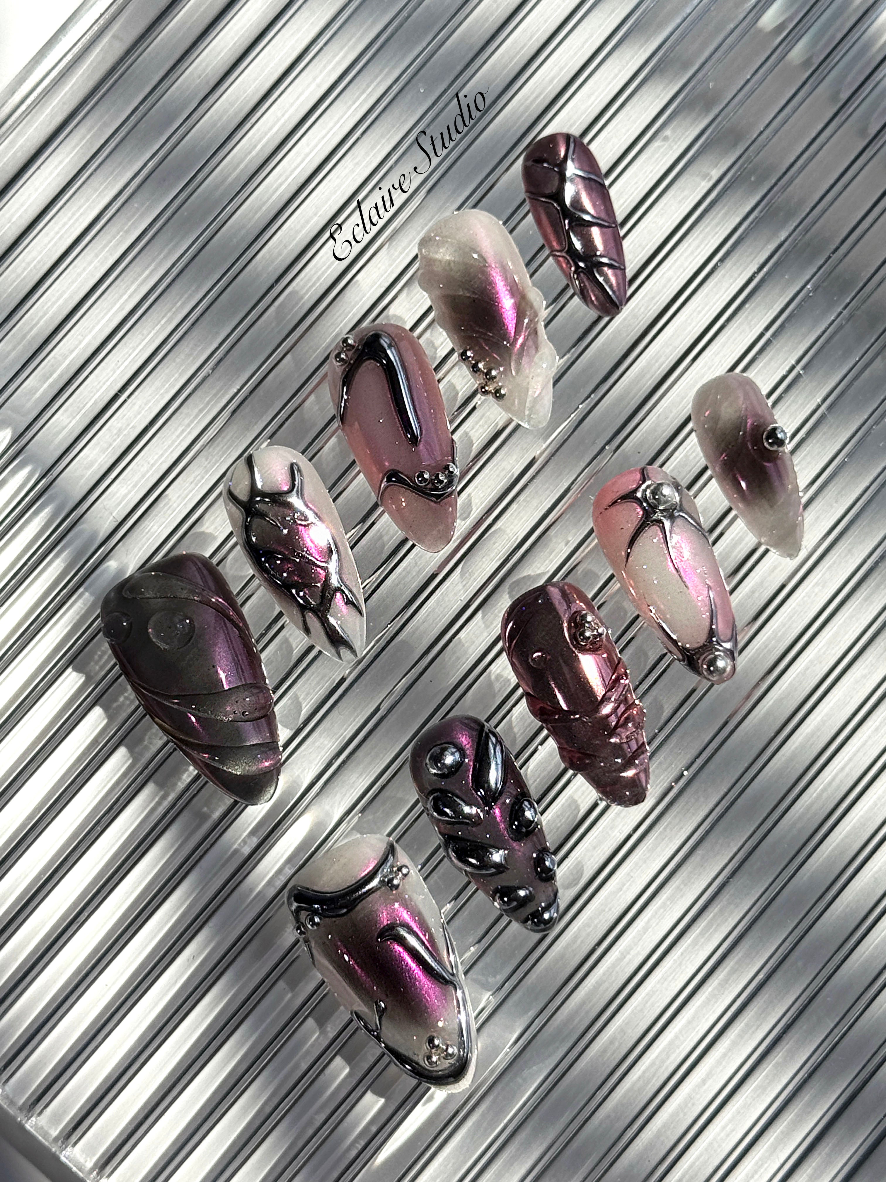 Set of long, almond-shaped press-on nails with a pink-to-purple multichrome base, accented by sculpted chrome designs, metallic textures, and small silver studs, displayed on a ribbed reflective surface under natural light.