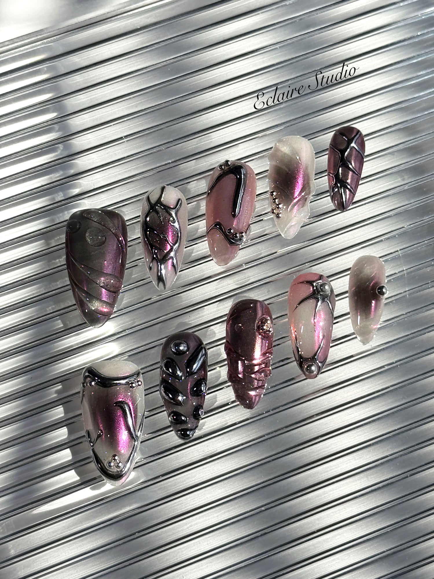 Set of long, almond-shaped press-on nails with a pink-to-purple multichrome base, accented by sculpted chrome designs, metallic textures, and small silver studs, displayed on a ribbed reflective surface under natural light.