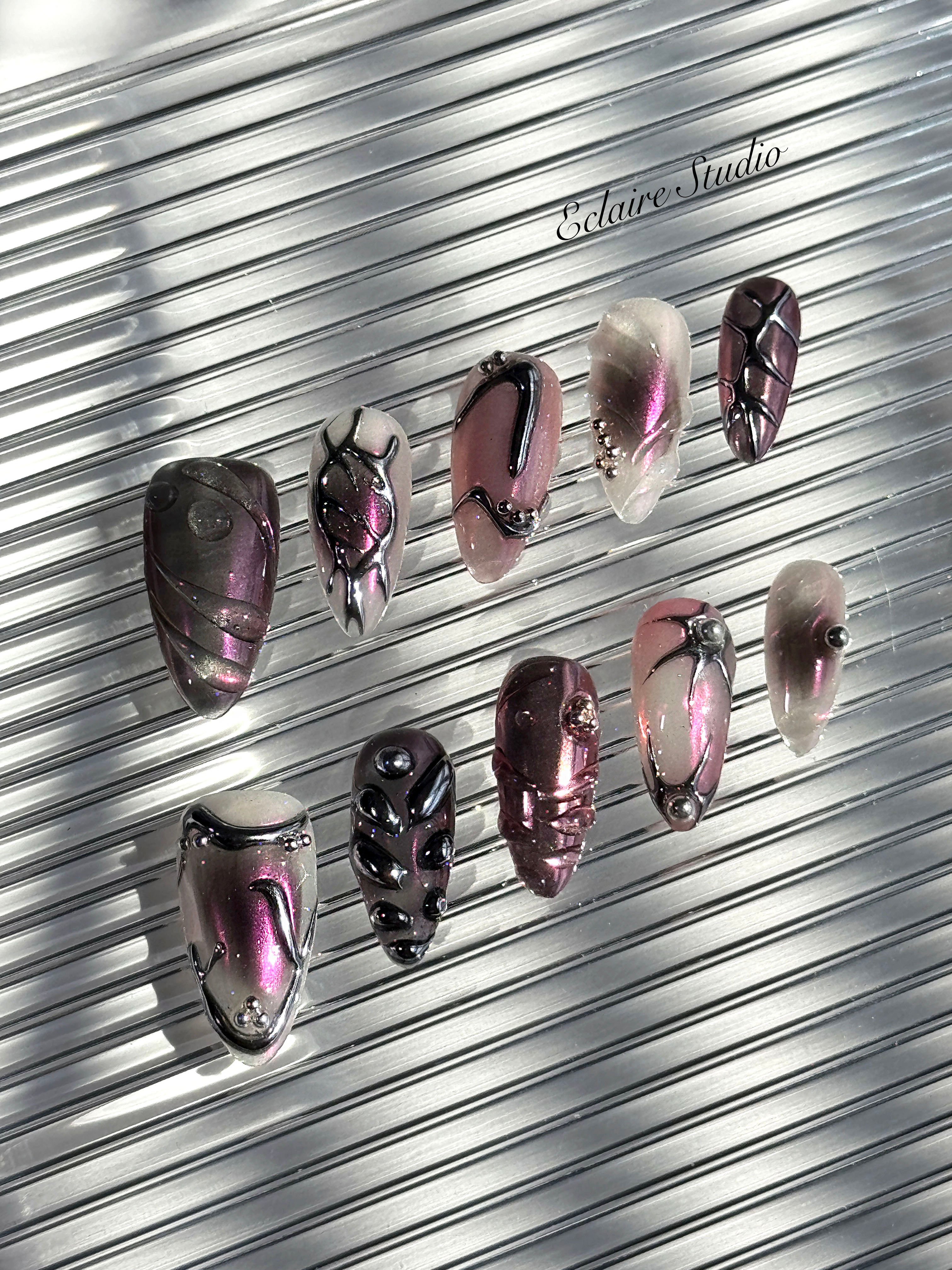 Set of long, almond-shaped press-on nails with a pink-to-purple multichrome base, accented by sculpted chrome designs, metallic textures, and small silver studs, displayed on a ribbed reflective surface under natural light.